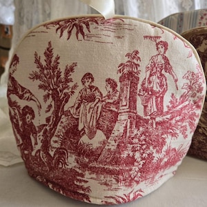 May include: A red and white toile tea cozy with a ribbon handle. The design features a pastoral scene with people and trees.
