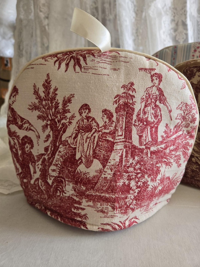 May include: A red and white toile tea cozy with a ribbon handle. The design features a pastoral scene with people and trees.