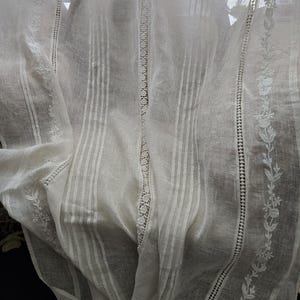 May include: A white, sheer curtain with vertical stripes and floral embroidery. The curtain features delicate lace detailing down the centre and along the edges. The fabric is translucent, allowing light to pass through, creating a soft, airy feel.