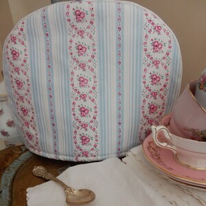 May include: A white teapot cozy with blue and pink floral stripes. The cozy is sitting on a table with a silver spoon and a pink teacup and saucer.