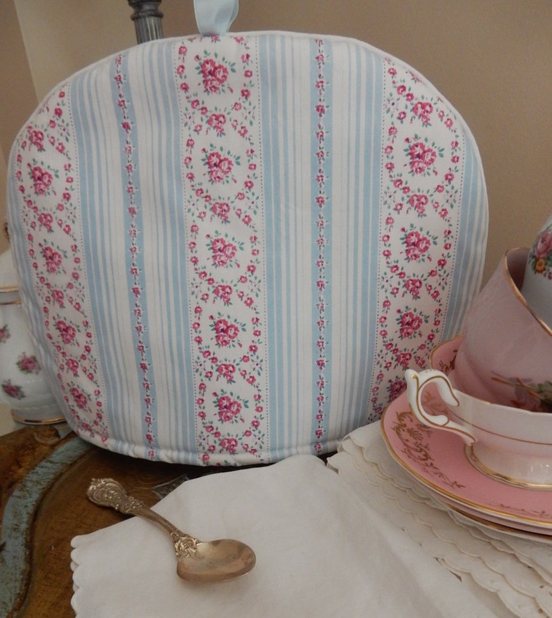 May include: A white teapot cozy with blue and pink floral stripes. The cozy is sitting on a table with a silver spoon and a pink teacup and saucer.