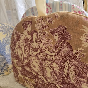 May include: A brown and beige tea cozy with a red and white toile pattern featuring three figures in a garden setting. The cozy has a cream-colored loop handle.