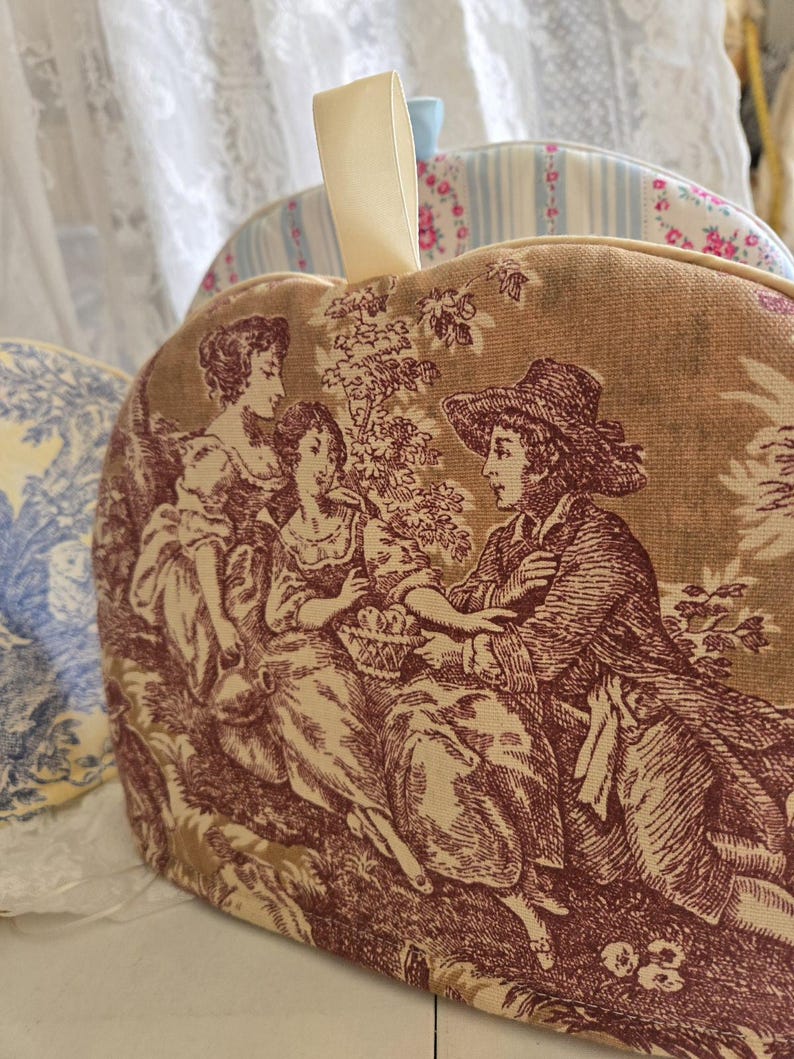 May include: A brown and beige tea cozy with a red and white toile pattern featuring three figures in a garden setting. The cozy has a cream-colored loop handle.