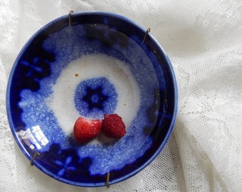 May include: A blue and white ceramic bowl with a floral pattern and two red strawberries inside. The bowl has a wire rim and is sitting on a white lace tablecloth.