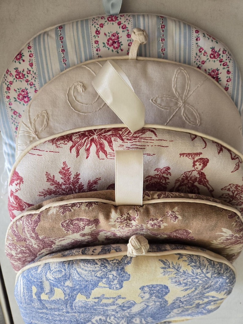 May include: Set of five cream-colored fabric pot holders with different patterns. Each pot holder has a cream-colored fabric loop for hanging. The patterns include a blue and white striped pattern, a cream-colored pattern with embroidered swirls, a red and white floral pattern, a brown and white floral pattern, and a blue and white floral pattern.