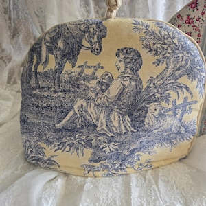 May include: A yellow tea cozy with a blue and white toile pattern featuring a man sitting with a dog and a horse in the background.