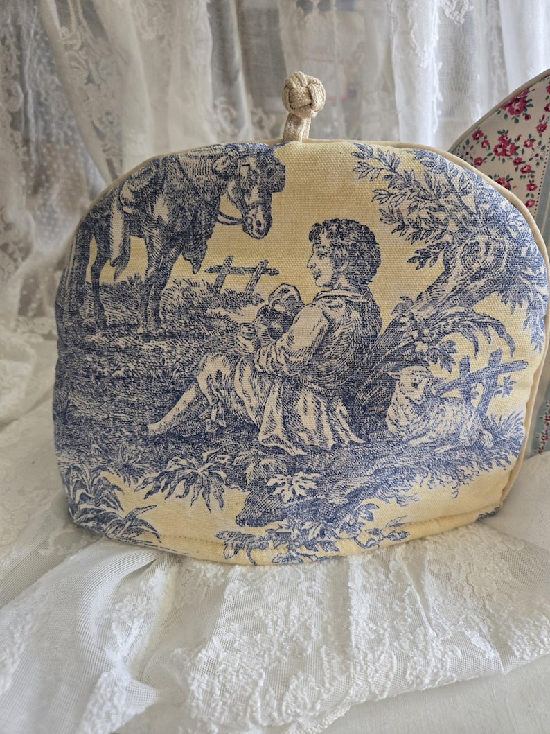 May include: A yellow tea cozy with a blue and white toile pattern featuring a man sitting with a dog and a horse in the background.