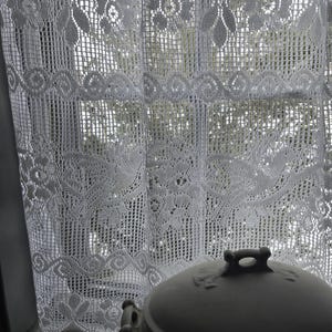 Peut inclure: Un rideau blanc en dentelle avec un motif floral est suspendu devant une fenêtre. Un plat en céramique blanc de forme ovale avec un couvercle est posé sur le rebord de la fenêtre. Le plat a une poignée sur le couvercle et deux poignées latérales.