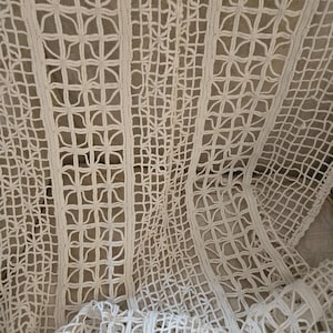 May include: Close-up of a cream-colored, open-weave textile. The fabric features a repeating pattern of squares and floral-like shapes, creating a delicate, lacy appearance. The texture is soft and the material drapes gently, suggesting a decorative item.