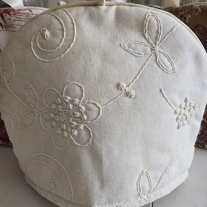 May include: A white cotton tea cozy with embroidered floral designs. The cozy has a round shape and a drawstring closure.