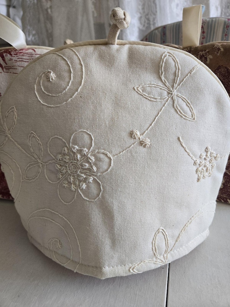 May include: A white cotton tea cozy with embroidered floral designs. The cozy has a round shape and a drawstring closure.