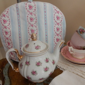 May include: A white teapot with a floral pattern and gold trim, sitting on a gold tray with a tea cozy in the background. The tea cozy is blue and white with a floral pattern.