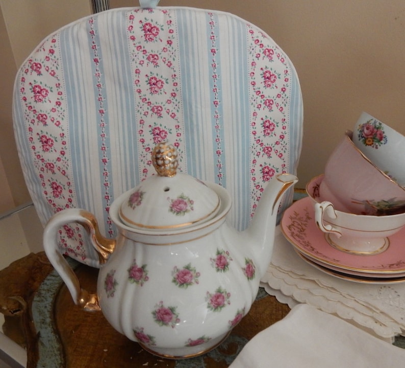 May include: A white teapot with a floral pattern and gold trim, sitting on a gold tray with a tea cozy in the background. The tea cozy is blue and white with a floral pattern.