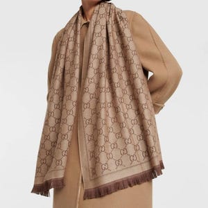 May include: A rectangular scarf in a light brown and dark brown colour scheme featuring a repeating interlocking GG logo pattern throughout the fabric. The scarf has a finished border and short, straight fringe detailing along the bottom edges.