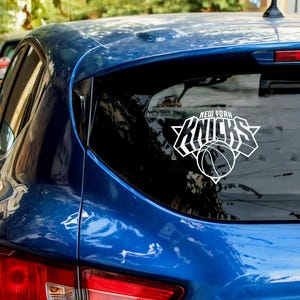 May include: A blue car with a white New York Knicks logo sticker on the rear window. The sticker features the team name and a basketball graphic. The car is parked outdoors, with trees and other vehicles visible in the background.
