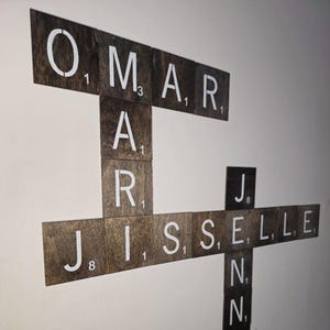 May include: Wooden Scrabble tile wall art spelling out "OMAR", "AR", "JISSELLE", and "NNY" in white letters. The tiles are arranged in a crossword style, creating a personalized decorative piece.