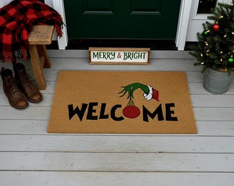 May include: A rectangular coir doormat with the word "WELCOME" in black block letters. The "O" is replaced with a red ornament held by a green hand with a white cuff and red sleeve. A small Christmas tree and a "Merry & Bright" sign are also visible.