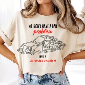 Puede incluir: Camiseta color crema con el texto "NO I DON'T HAVE A CAR problem _ I HAVE A ASTORAGE PROBLEM" en negro y rojo. La camiseta presenta un dibujo lineal de un coche de carreras.