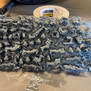 May include: A collection of small, gray plastic model parts, possibly for miniatures or a similar hobby, are arranged in a rectangular shape. The parts are embedded in a clear, viscous substance, likely resin, and are partially visible.