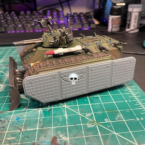 May include: A miniature olive green military tank model featuring a front-mounted plow, a turret with a cannon, and a white missile on the side. The tank is equipped with grey side armor panels decorated with a white skull and wing emblem.