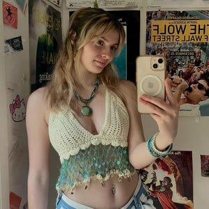 Può includere: Top all'uncinetto color crema con scollo a V profondo e rifiniture con paillettes e frange turchesi. Abbinato a pantaloncini a righe blu e bianche. Accessori: collana e braccialetti turchesi.
