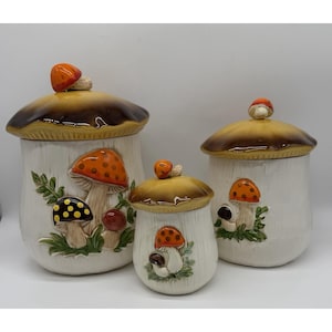 May include: Set of three ceramic mushroom-shaped canisters. The canisters are white with brown and orange mushroom cap lids. Each canister features a different mushroom design with green foliage accents. The largest canister is approximately 10 inches tall.