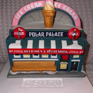 Könnte beinhalten: Ein Keramikmodell eines alten Eiscafés namens "Polar Palace". Das Geschäft hat eine grün-weiß gestreifte Markise mit einem Schild, auf dem "5¢ Coca-Cola DELICIOUS & REFRESHING Coca-Cola 5¢" steht. Vor dem Geschäft befindet sich eine Holzbank und an der Seite eine Coca-Cola-Flasche.