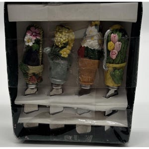 May include: A set of four decorative bottle stoppers, each featuring a different floral design. The stoppers are in a black box and include a basket of flowers, a watering can, a terracotta pot, and a yellow pot with tulips.