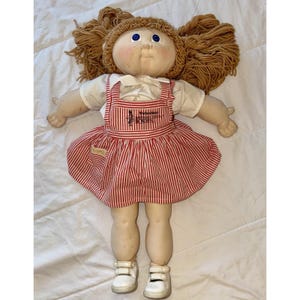 May include: A vintage doll with a fabric body, tan yarn hair, and blue eyes. The doll wears a white shirt under a red and white striped dress with the text "Original Cabbage Patch Kids" on the front. White shoes complete the outfit.