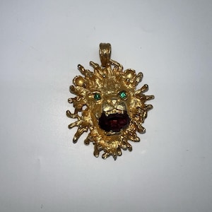 May include: A gold-colored lion pendant with a textured mane and face. The lion has green eyes and a dark red gemstone for its mouth. The pendant is designed with a loop at the top for a chain.