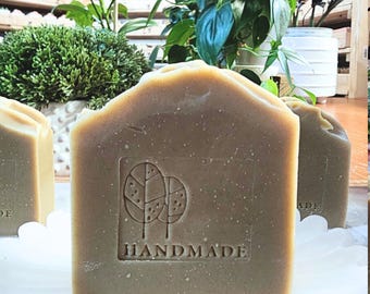 May include: Close-up of handmade soap bars with a light brown color. The soap has a tree design and the word "HANDMADE" embossed on the front. The soap is on a white, scalloped edge plate. Green plants are in the background.