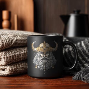 May include: A black ceramic mug featuring a detailed illustration of a Viking warrior with a golden horned helmet and a long, white beard. The mug has a large handle and is set against a cozy, dark background.