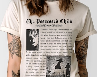 Op de afbeelding: Crèmekleurig T-shirt met de tekst "The Possessed Child" in een gotisch lettertype. Het shirt bevat een zwart-wit afbeelding van een meisje en een scène met twee figuren. De tekst beschrijft het verhaal van een jong meisje.