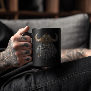 May include: Black ceramic mug featuring a detailed illustration of a Viking warrior with a horned helmet and long beard. The Viking's helmet is gold and the beard is gray and white. The mug has a black handle.
