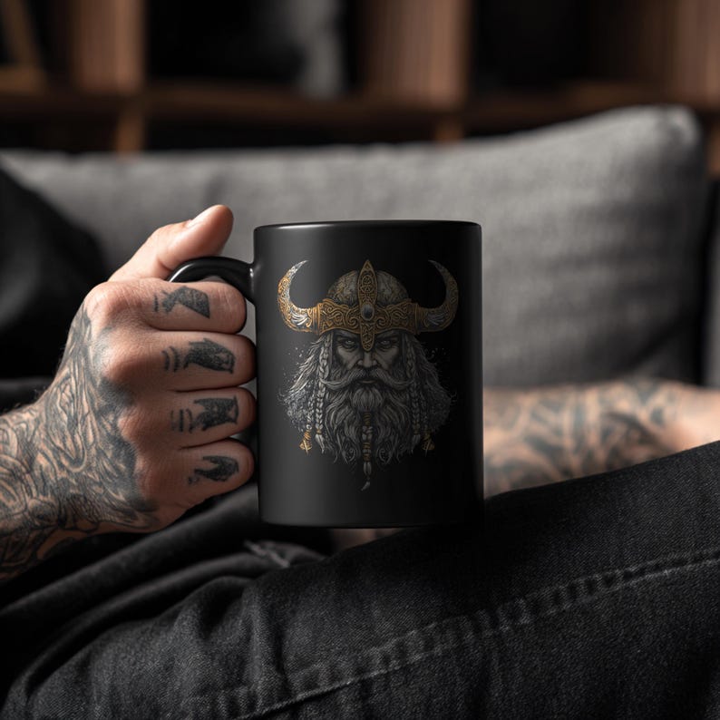 May include: Black ceramic mug featuring a detailed illustration of a Viking warrior with a horned helmet and long beard. The Viking's helmet is gold and the beard is gray and white. The mug has a black handle.