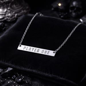 Op de afbeelding: Zilverkleurige ketting met een rechthoekige hanger met de tekst "PLAYER ONE". De fijne ketting en hanger liggen op een zwarte fluwelen ondergrond.