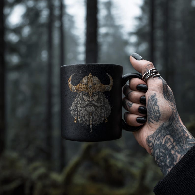 May include: Black ceramic mug featuring a detailed illustration of a Viking warrior with a horned helmet and long beard. The artwork is in shades of gold and brown. The mug is held against a blurred forest background.