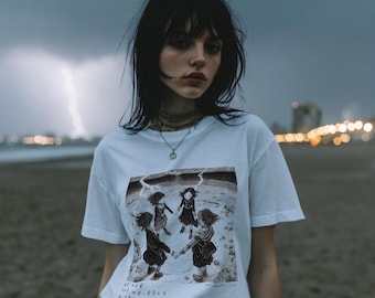 Könnte beinhalten: Weißes T-Shirt mit einer Schwarz-Weiß-Illustration von vier Mädchen, die sich an den Händen halten. Auf der Vorderseite steht der Text "WE ARE THE WEIRD SISTER". Die Person steht am Strand.