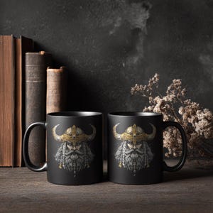 May include: Two black ceramic mugs with a detailed illustration of a Viking warrior. The warrior has a long white beard, a golden helmet with horns, and a stern expression. The mugs are set on a wooden surface with books in the background.