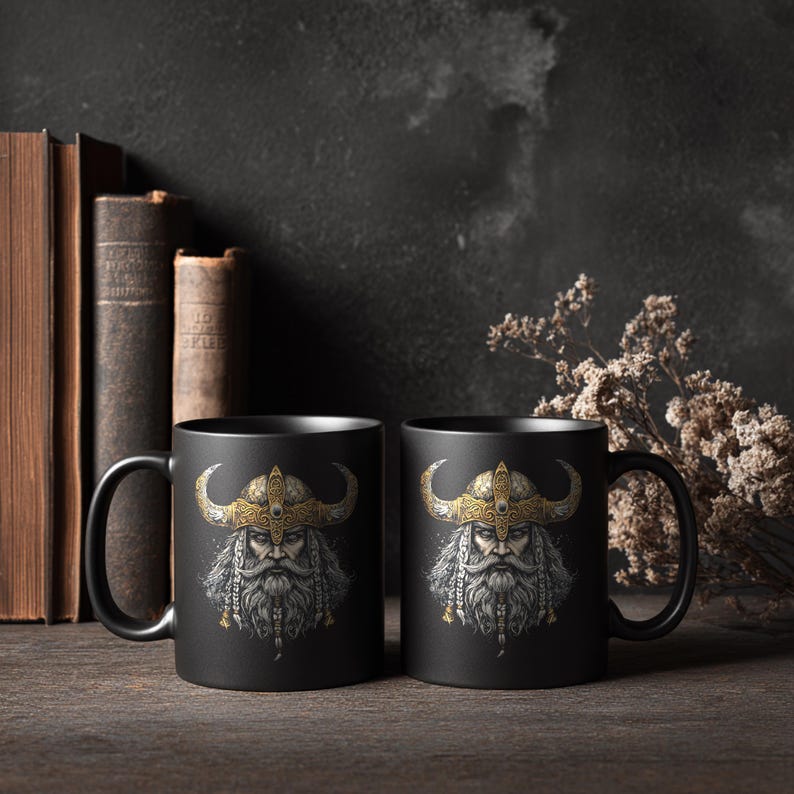 May include: Two black ceramic mugs with a detailed illustration of a Viking warrior. The warrior has a long white beard, a golden helmet with horns, and a stern expression. The mugs are set on a wooden surface with books in the background.