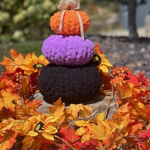 Op de afbeelding: Een decoratieve herfststapel van drie pluche pompoenen in oranje, paars en zwart. De bovenste pompoen heeft een bruin lint. De pompoenen zijn omringd door kleurrijke herfstbladeren en zonnebloemen, wat een feestelijke seizoensweergave creëert.