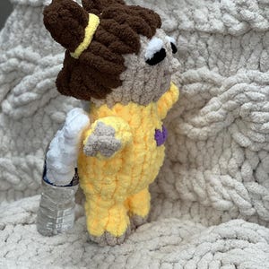 K&ouml;nnte beinhalten: Ein geh&auml;keltes Pl&uuml;schtier in Form eines M&auml;dchens, das ein gelbes und violettes Outfit tr&auml;gt. Das Spielzeug hat braunes Haar und gro&szlig;e schwarze Augen.