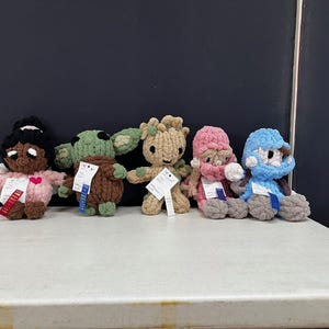 May include: Five handmade crochet dolls are displayed on a white surface. The dolls include a girl with a pink dress, a green alien, a brown tree-like figure, a pink-hatted figure, and a blue-hatted figure. Each doll has a tag attached.