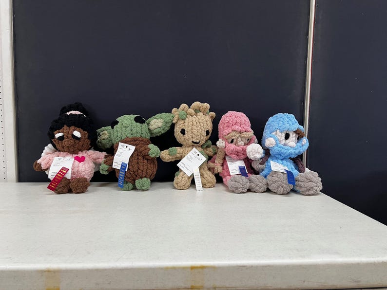 May include: Five handmade crochet dolls are displayed on a white surface. The dolls include a girl with a pink dress, a green alien, a brown tree-like figure, a pink-hatted figure, and a blue-hatted figure. Each doll has a tag attached.