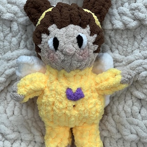 K&ouml;nnte beinhalten: Ein geh&auml;keltes Pl&uuml;schtier in Form eines M&auml;dchens mit braunen Haaren, das ein gelbes Outfit mit einem lila Herz auf der Brust tr&auml;gt. Das Spielzeug hat Fl&uuml;gel und sitzt auf einer wei&szlig;en flauschigen Decke.