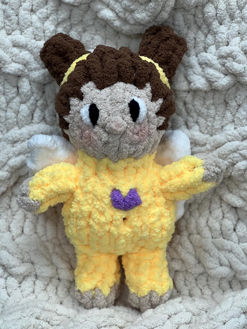K&ouml;nnte beinhalten: Ein geh&auml;keltes Pl&uuml;schtier in Form eines M&auml;dchens mit braunen Haaren, das ein gelbes Outfit mit einem lila Herz auf der Brust tr&auml;gt. Das Spielzeug hat Fl&uuml;gel und sitzt auf einer wei&szlig;en flauschigen Decke.