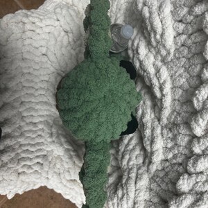May include: A handmade green plush toy with black accents, possibly a creature or character, rests on a thick, white, chunky knit blanket. A clear plastic water bottle is partially visible near the toy. The blanket has a soft, textured appearance.