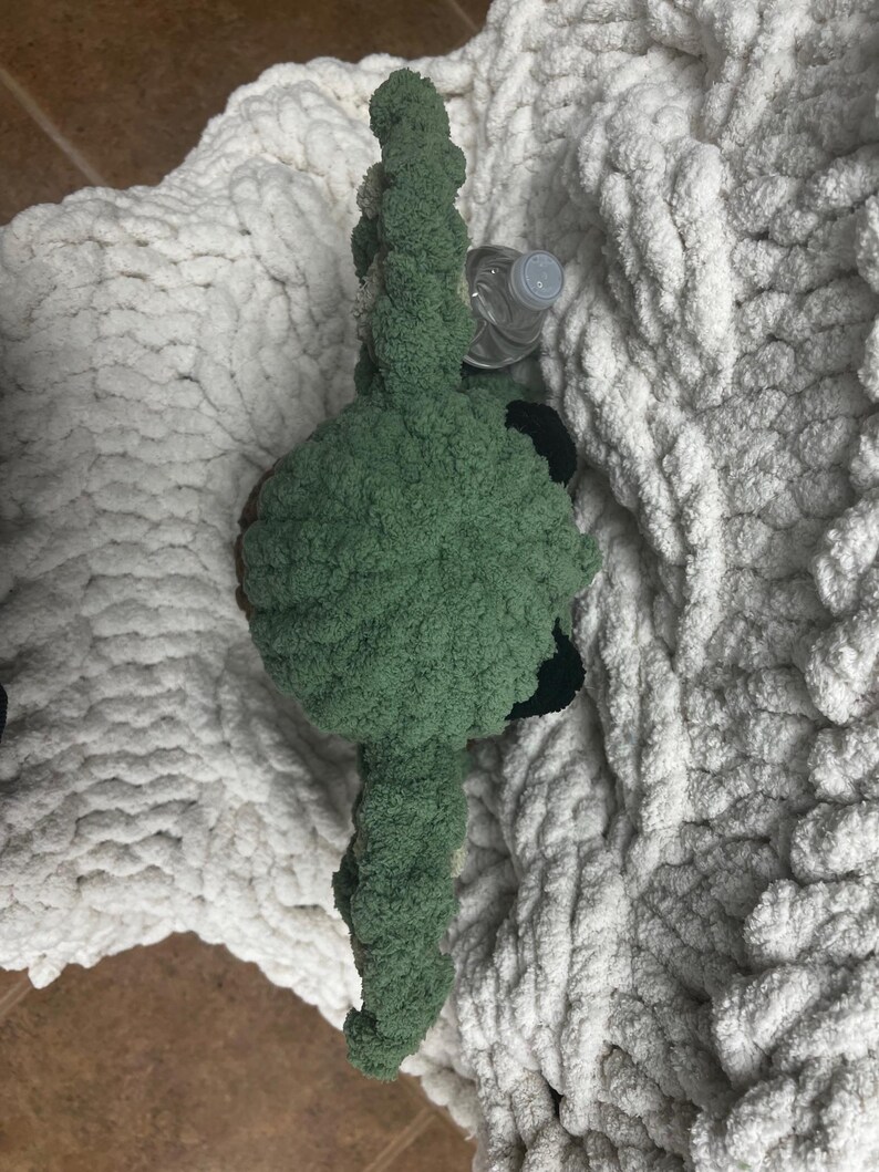 May include: A handmade green plush toy with black accents, possibly a creature or character, rests on a thick, white, chunky knit blanket. A clear plastic water bottle is partially visible near the toy. The blanket has a soft, textured appearance.