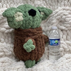 May include: A handmade crochet plush toy in the likeness of a character with large ears and a brown robe. The toy is green and brown, with black eyes. A clear plastic water bottle is next to the toy.