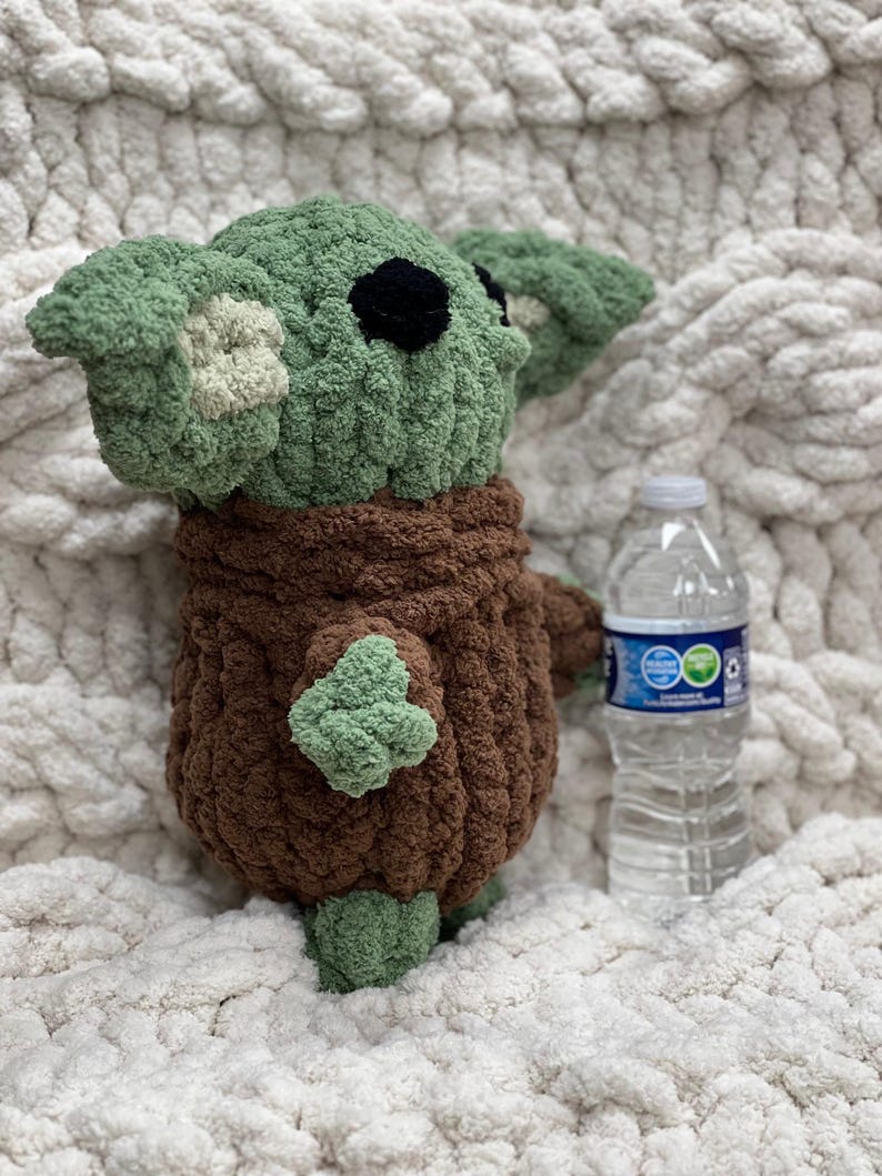 May include: A handmade crochet plush toy in the likeness of a character with large ears and a brown robe. The toy is green and brown, with black eyes. A clear plastic water bottle is next to the toy.